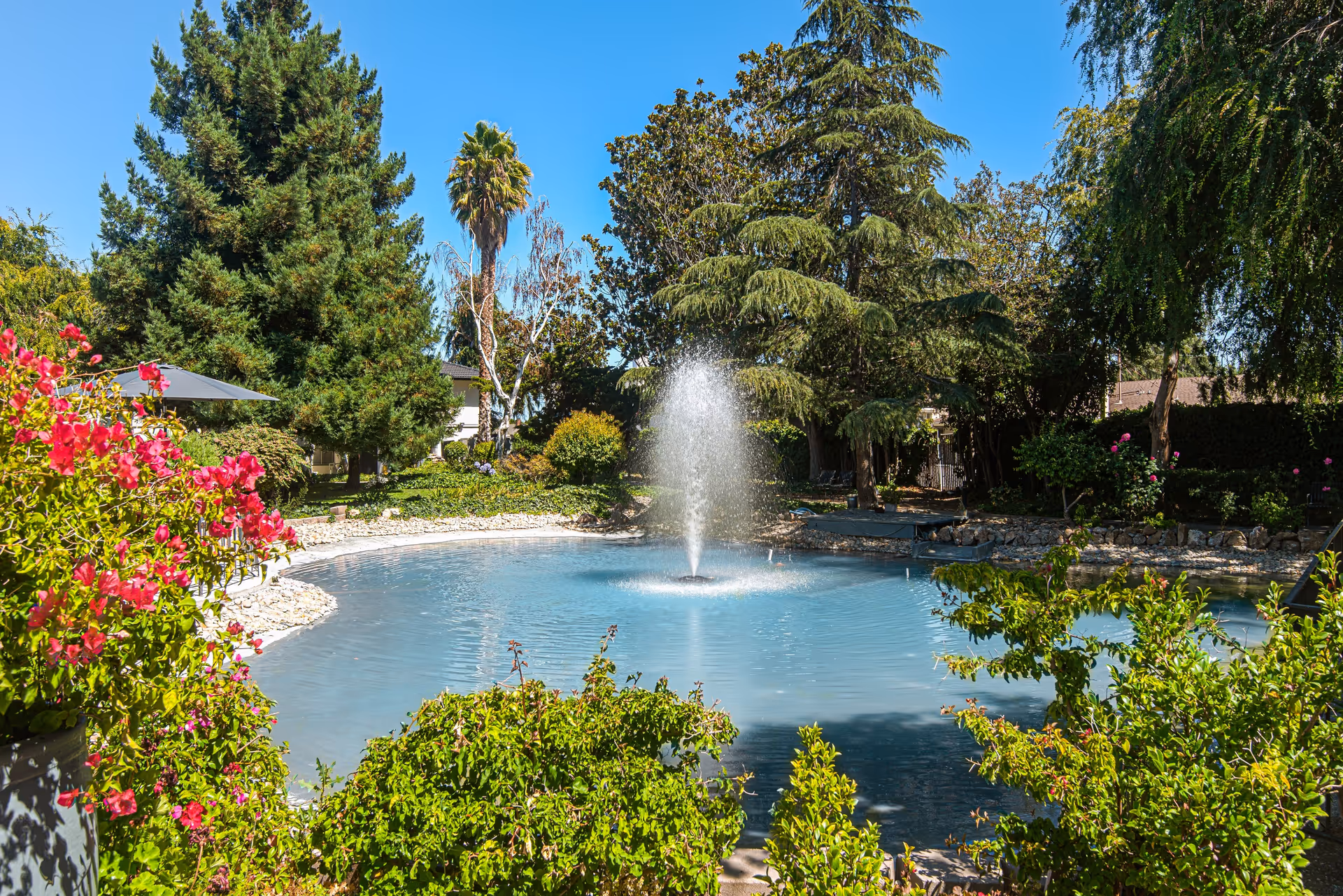A serene outdoor garden area at Villa Serena Retirement Community featuring a circular pond with a central water fountain surrounded by lush greenery, colorful flowers, and tall trees under a clear blue sky.