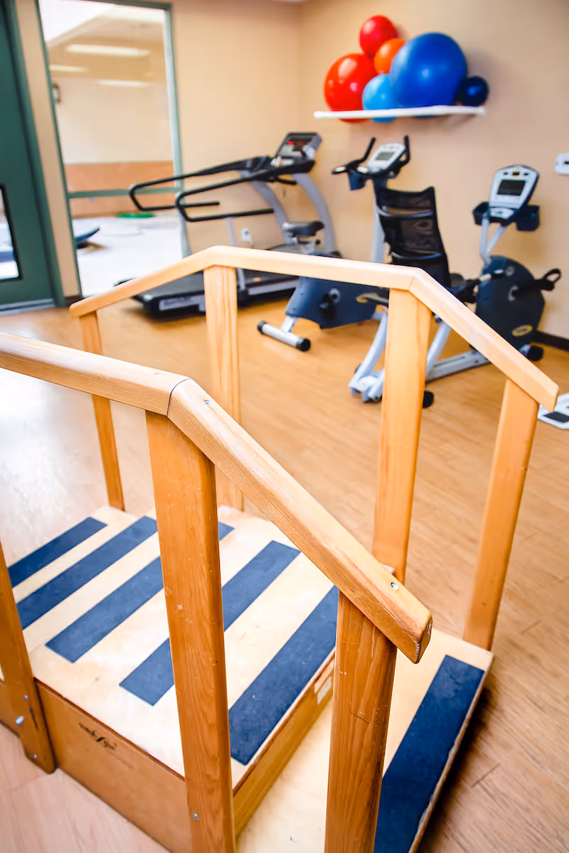 Indoor exercise room with a wooden rehabilitation step platform in the foreground and exercise bikes and a treadmill in the background. Colorful exercise balls are placed on a wall shelf.