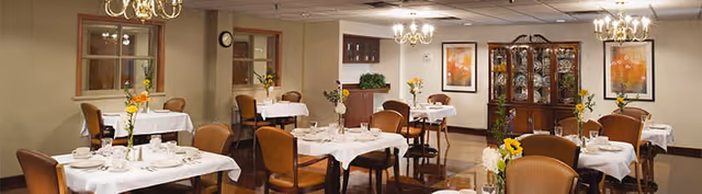 A dining room with several tables covered in white tablecloths, each set with plates, glasses, and silverware. The room features wooden chairs, chandeliers, framed artwork on the walls, and a china cabinet displaying dishes. Fresh flowers are placed on each table and around the room.