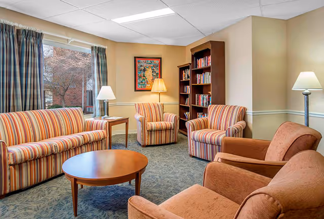 A cozy living room area with a striped sofa, two matching striped armchairs, and two brown armchairs arranged around a round wooden coffee table. There are two lamps providing warm lighting, a wooden bookshelf filled with books, a framed artwork on the wall, and windows with plaid curtains showing a view of trees and a brick building outside.