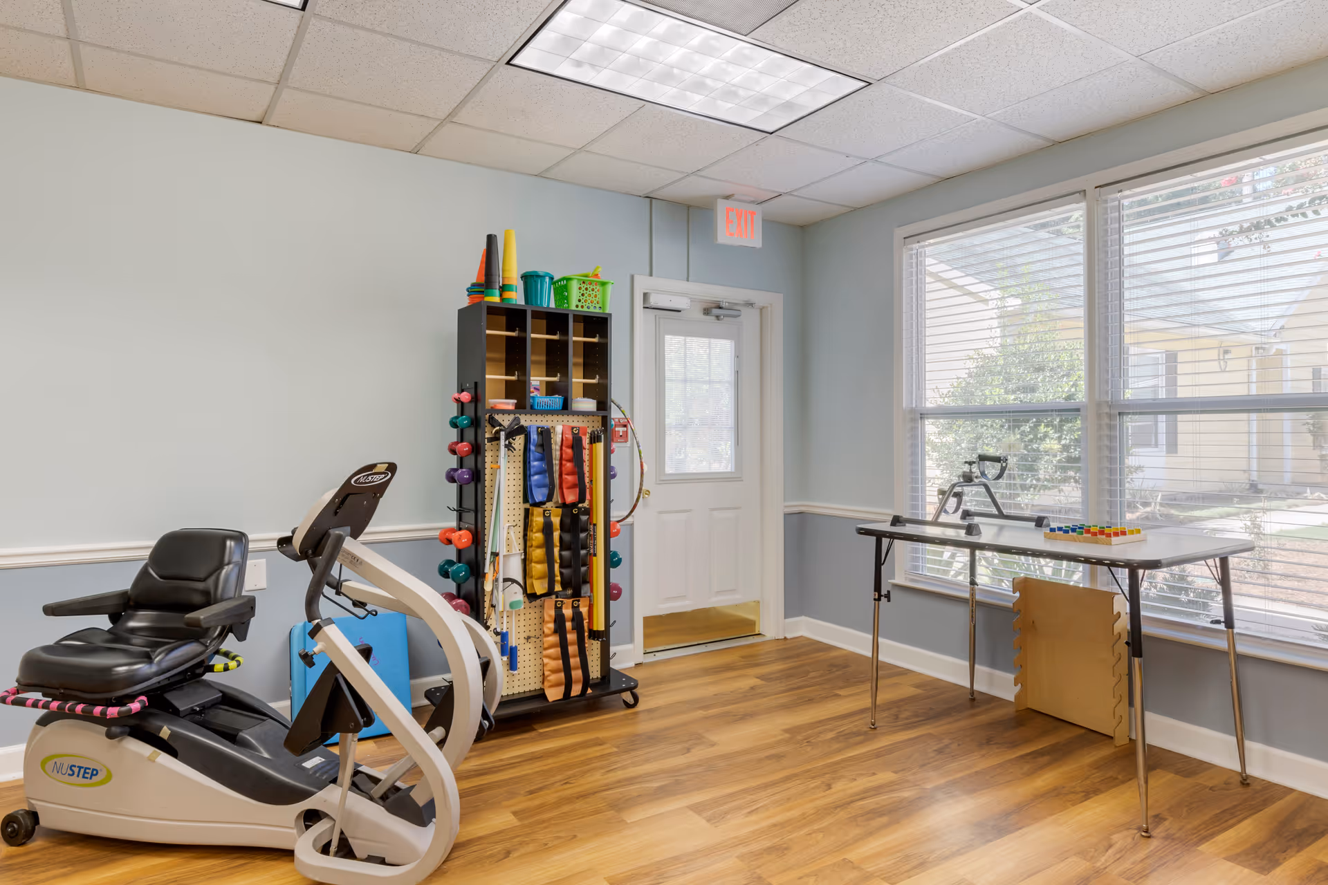 A small exercise room with a recumbent exercise bike, a rack holding various exercise equipment including dumbbells and resistance bands, a table with a hand exercise device and colorful blocks, and large windows letting in natural light.
