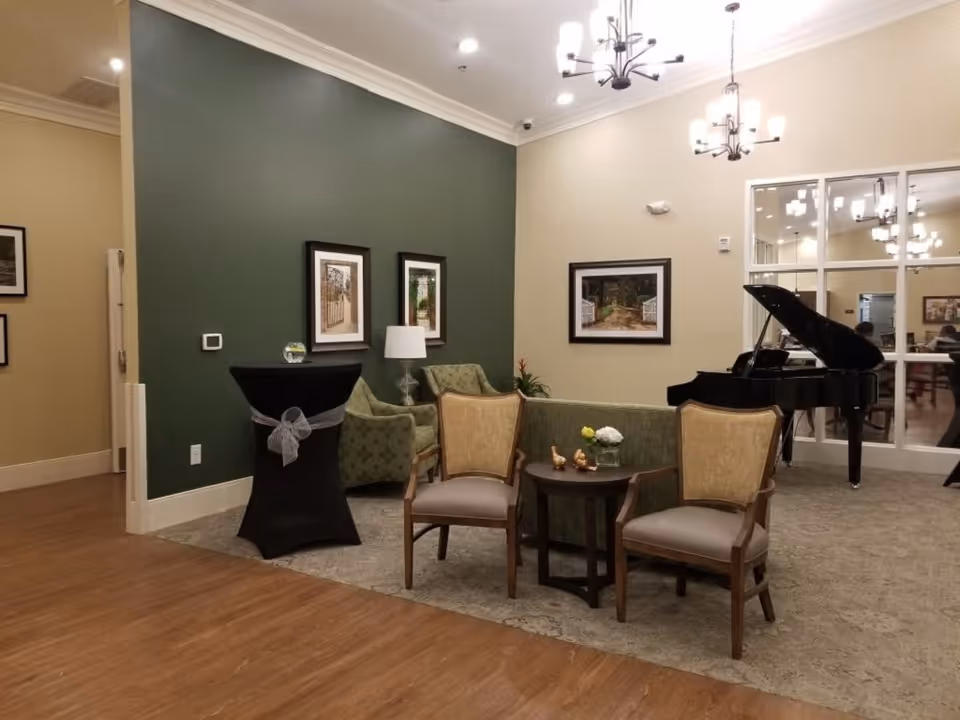 A furnished lounge area with chairs, small tables, framed artwork on a green accent wall and a grand piano.