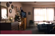 Sunlit sitting and dining area with a table and chairs, dresser with decorative items and a wall-mounted TV beneath a curtained window.
