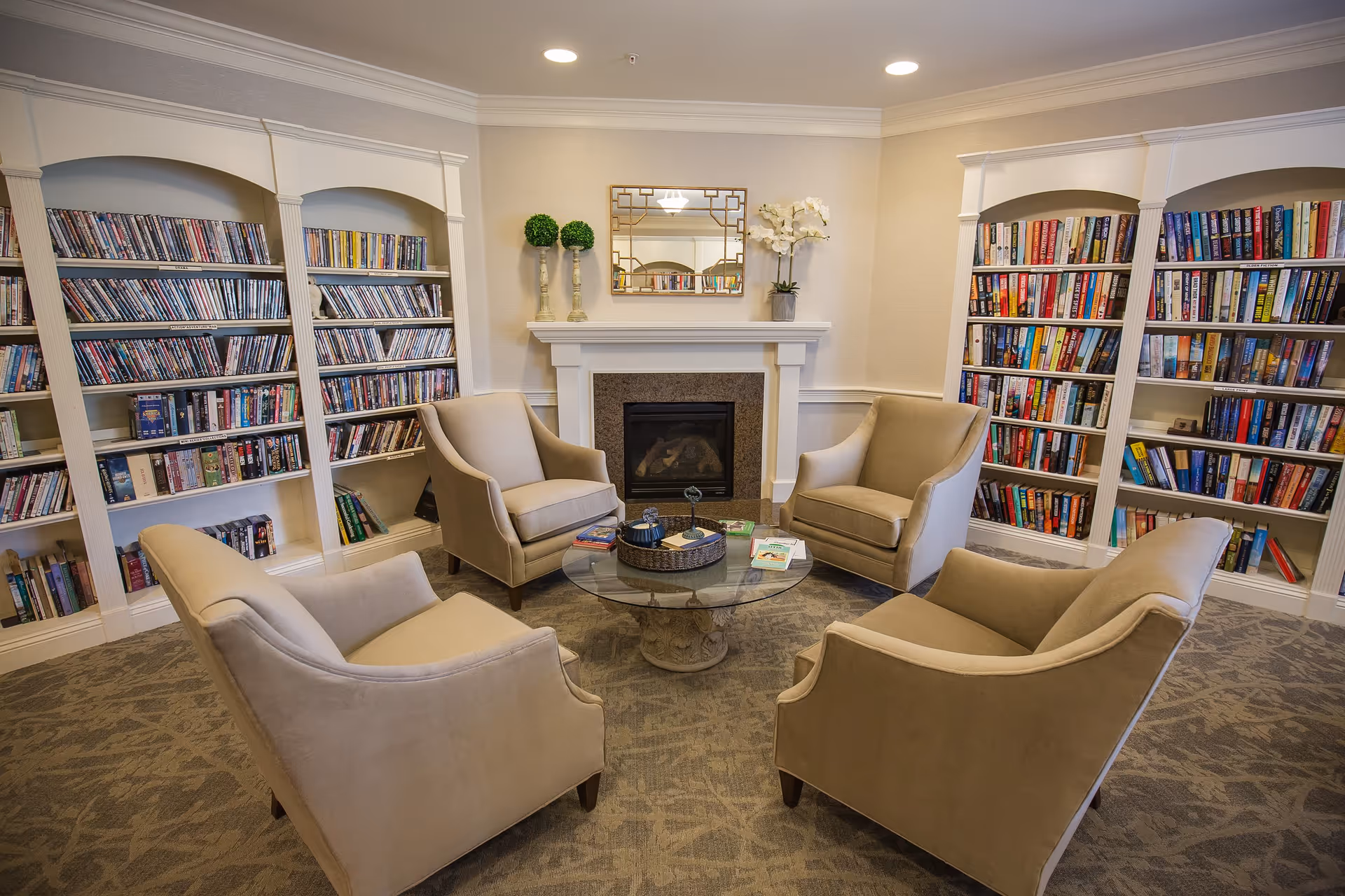 A cozy reading room with four beige armchairs arranged around a round glass coffee table. The room features built-in white bookshelves filled with books and DVDs on either side of a white fireplace with a decorative mirror and plants on the mantel. The carpet has a subtle pattern, and the ceiling has recessed lighting.