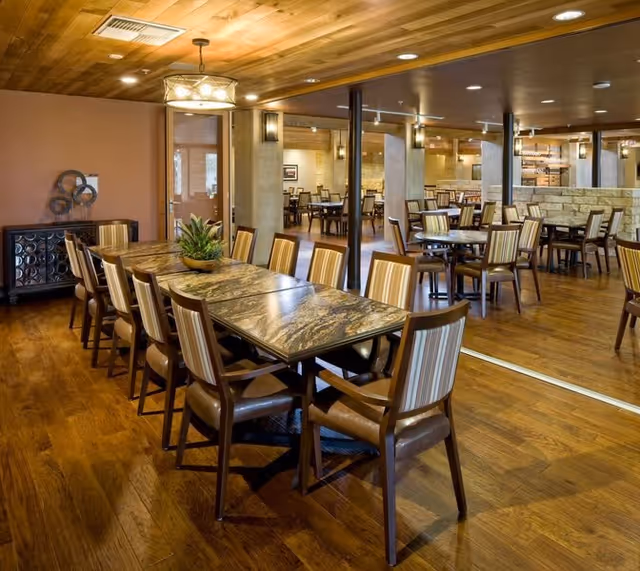 A spacious dining room with multiple tables and chairs arranged neatly. The room features wooden flooring and a wooden ceiling with recessed lighting and a hanging light fixture. There is a decorative plant centerpiece on the main table, and the walls have warm tones with some artwork and decor.