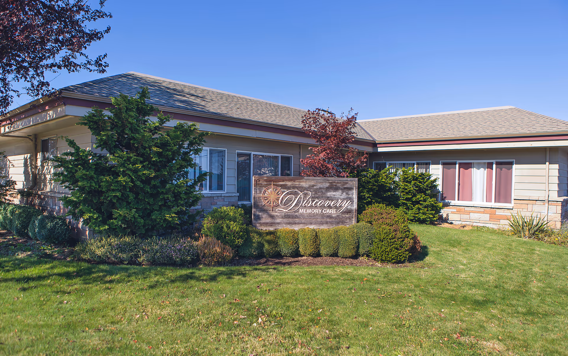 One-story care facility exterior with lawn, shrubs, and a wooden 'Discovery Memory Care' sign in front.