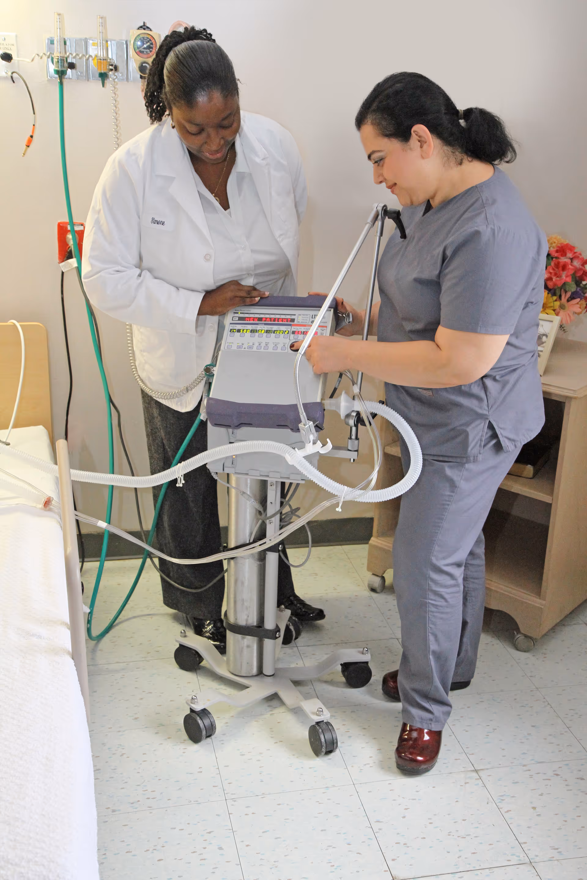 Two healthcare professionals, one in a white lab coat and the other in gray scrubs, are standing next to a medical ventilator machine in a clinical room with a hospital bed and medical equipment on the wall.