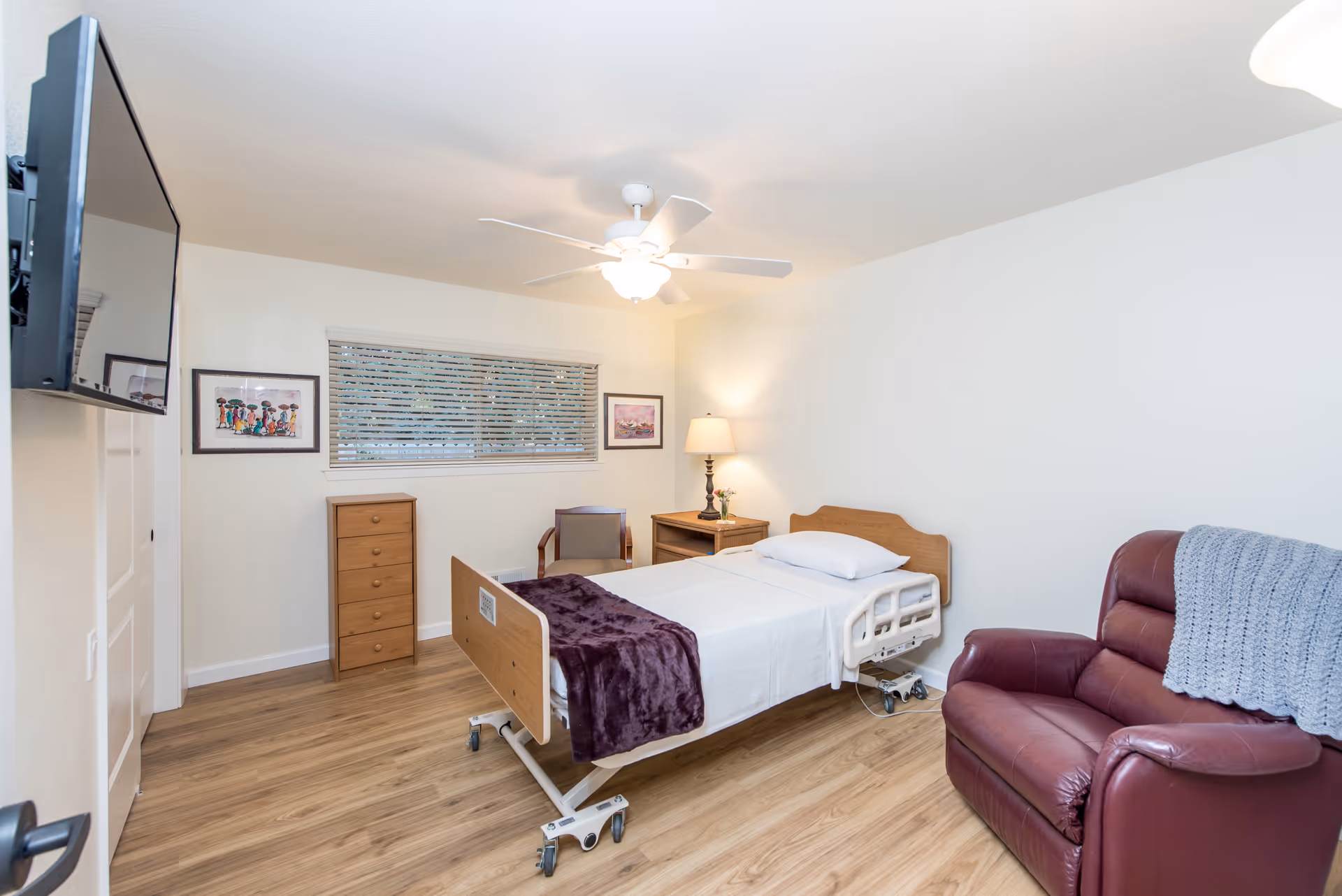 A simple, clean bedroom in a senior living facility with a single hospital-style bed covered with white sheets and a purple blanket. The room has light wood flooring, a maroon recliner chair with a gray knitted throw, a wooden nightstand with a lamp and flowers, a wooden chest of drawers, a chair, and two framed pictures on the wall. A ceiling fan with a light is on the ceiling, and a flat-screen TV is mounted on the wall opposite the bed.