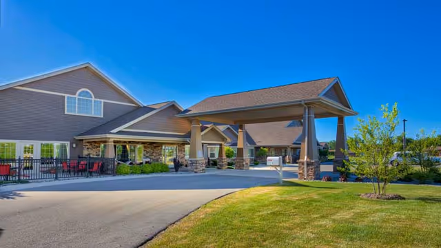 Exterior view of a senior living facility building with a covered entrance driveway, stone and siding facade, outdoor seating area with red chairs, green lawn, and clear blue sky.