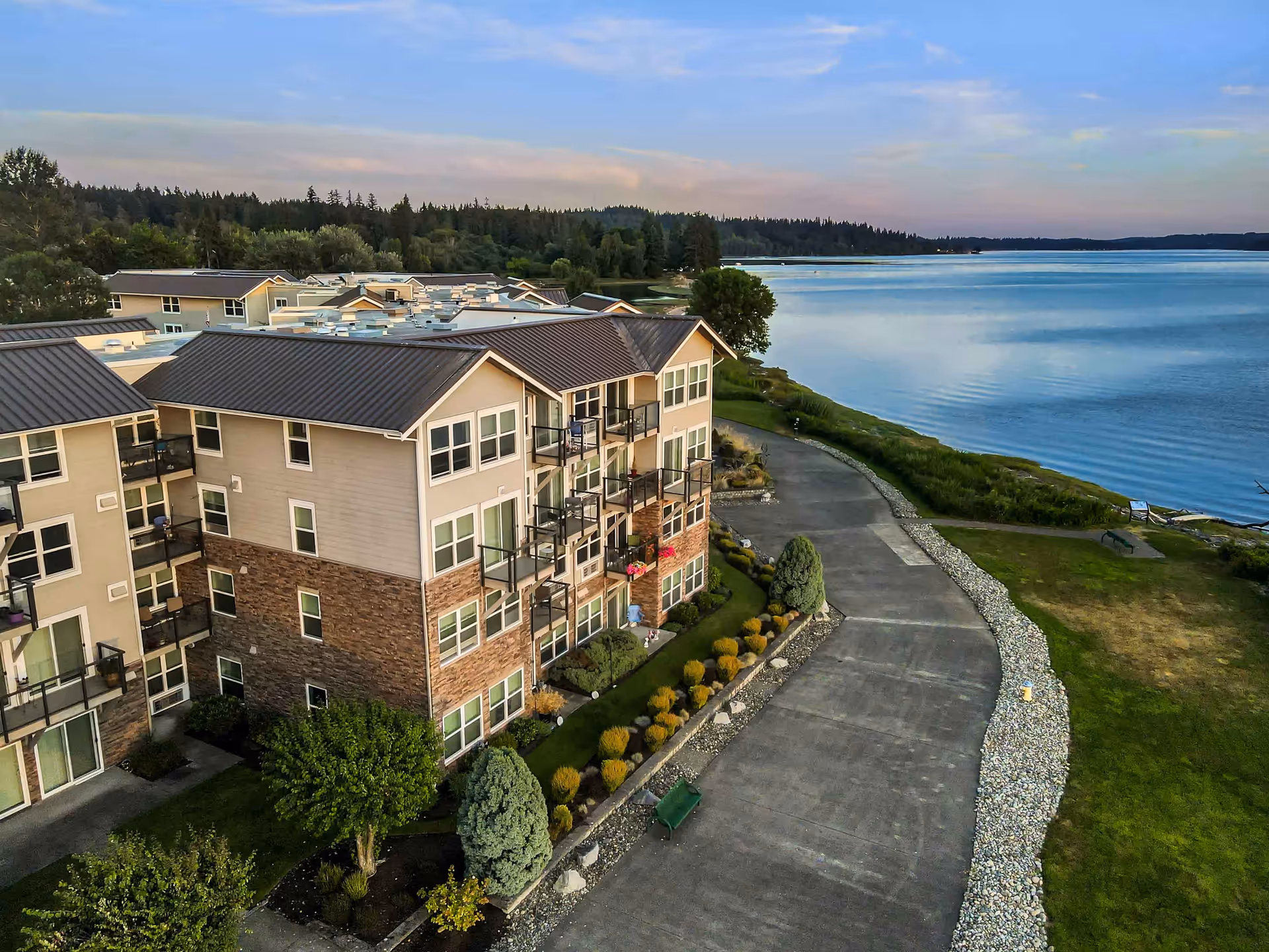 Aerial view of a multi-story residential building with balconies overlooking a large body of water. The building is surrounded by landscaped greenery, a paved walkway, and trees, with a forested area in the background under a partly cloudy sky.