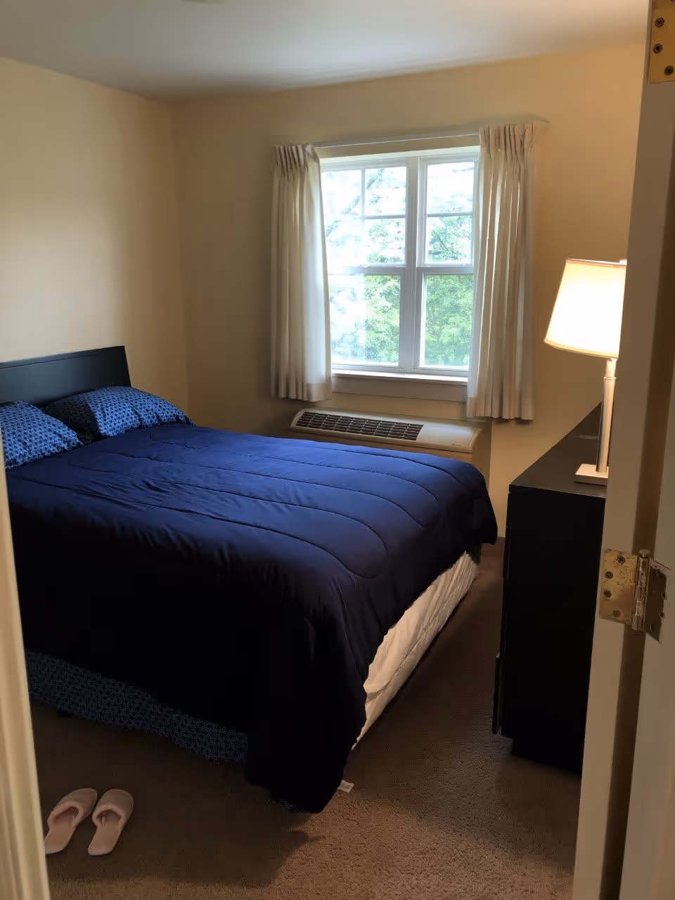 A small bedroom with a neatly made bed covered with a dark blue comforter and matching pillows. There is a window with white curtains letting in natural light, an air conditioning unit below the window, a black dresser with a lit lamp on top, and a pair of pink slippers on the carpeted floor near the bed.