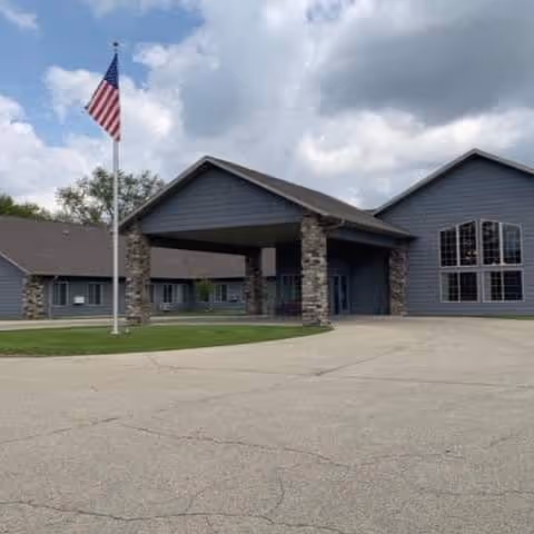 Exterior view of a single-story building with a covered entrance supported by stone pillars, an American flag on a flagpole in front, and a cloudy sky overhead.