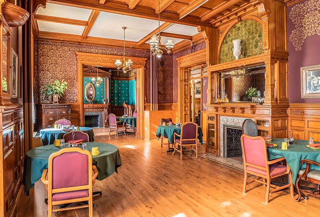 A warmly lit dining room with wooden paneling and ornate ceiling beams. Several round tables are covered with green tablecloths and surrounded by chairs with purple upholstery. The room features a decorative fireplace with a mirror above it, vintage-style chandeliers, and patterned wallpaper. A doorway leads to an adjoining room with similar decor and a fireplace.