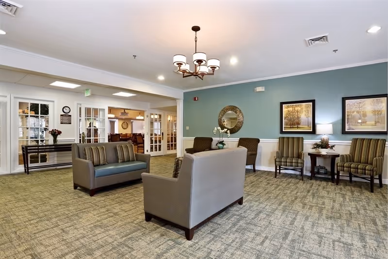 A spacious and well-lit common area in a senior living facility with two gray sofas facing each other, three striped armchairs along the wall, a round mirror, framed artwork, a small table with a lamp and plant, and a chandelier hanging from the ceiling. The walls are painted light blue with white trim, and there is a carpeted floor. In the background, glass doors lead to another room with more seating and a clock on the wall.