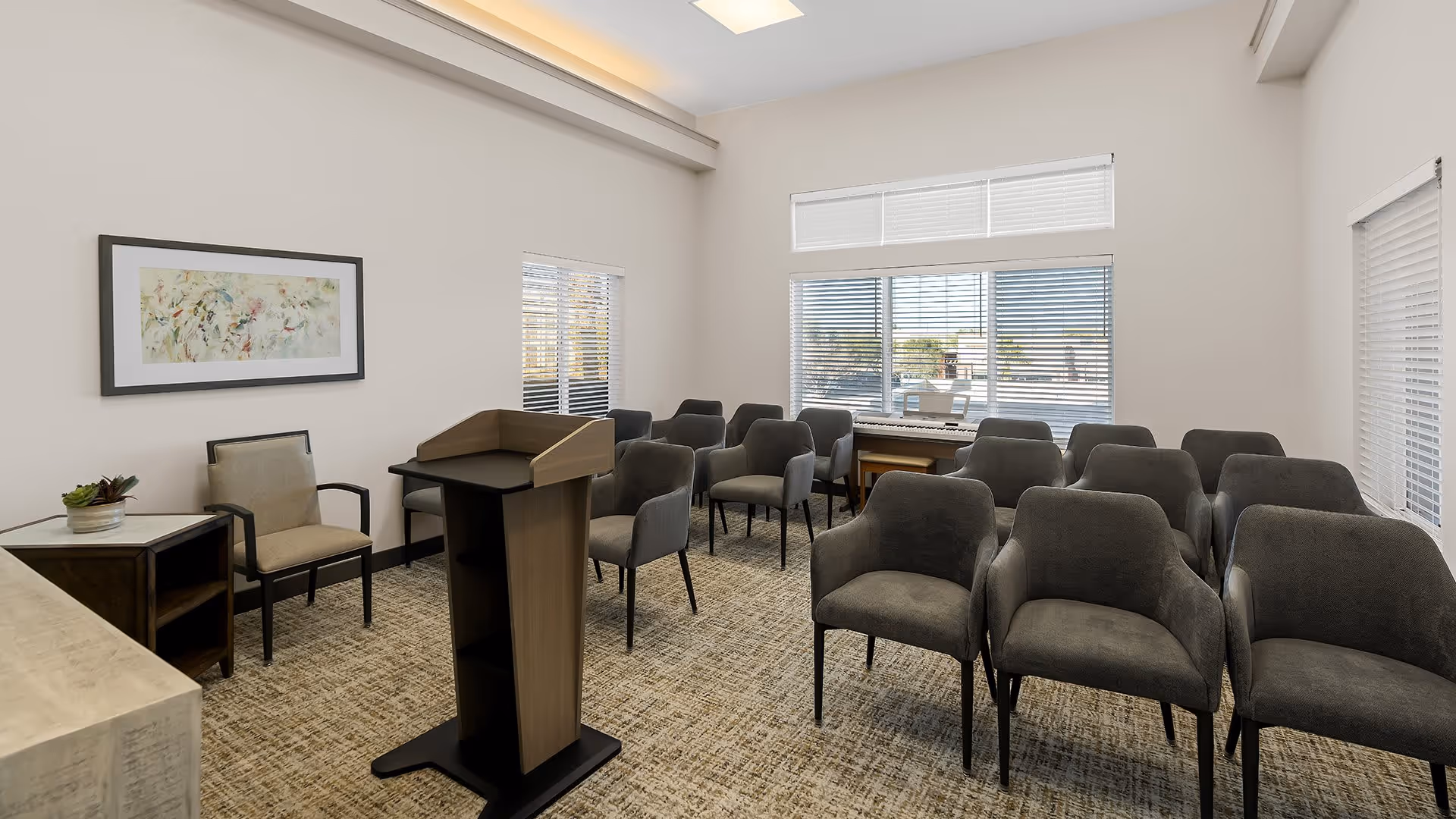 A small meeting or presentation room with several gray upholstered chairs arranged in rows facing a wooden podium. The room has beige walls, carpeted floor, and multiple windows with blinds allowing natural light. There is a framed abstract painting on one wall and a piano against the far wall under the windows.