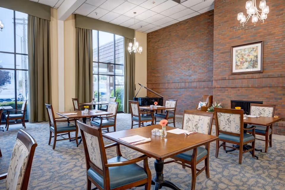 A bright dining room with several wooden tables and chairs arranged neatly. Each table has a small vase with a flower and some place settings. Large windows with green curtains allow natural light to fill the room. A brick wall with a framed painting and a chandelier hanging from the ceiling are visible. A black grand piano is placed against the brick wall.