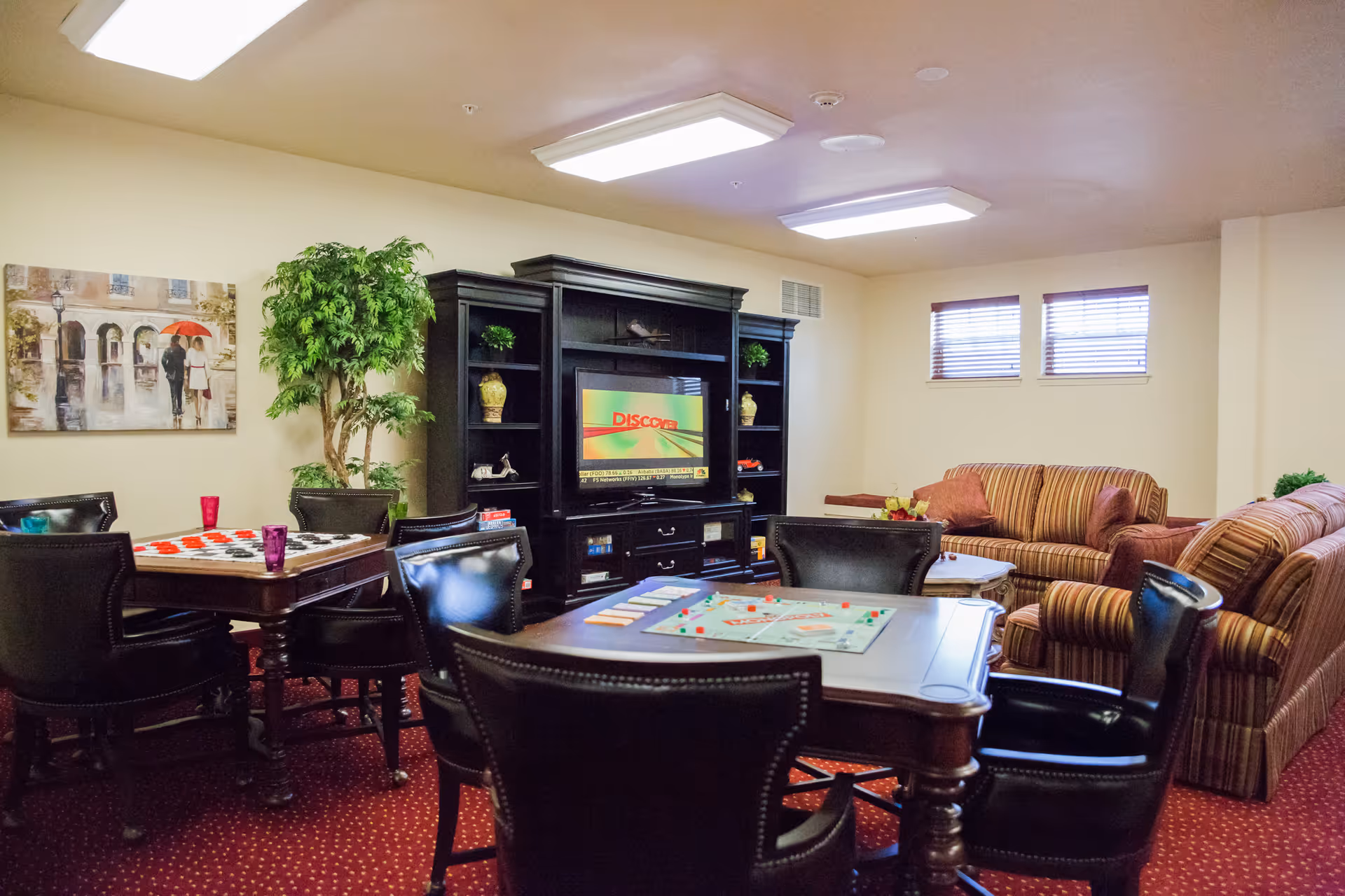 A cozy common room in a senior living facility with a red carpeted floor, two game tables with board games set up, black leather chairs around the tables, a large black entertainment center with a TV, a striped sofa and loveseat, a small coffee table, a large potted plant, and a painting on the wall.