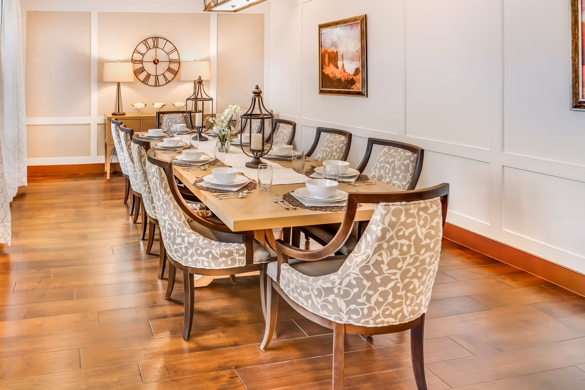 A well-lit dining room with a long wooden table set for eight people. The table is adorned with white plates, bowls, glasses, silverware, and decorative candle holders. The chairs have wooden frames with beige patterned upholstery. The room features wooden flooring, white paneled walls, a large wall clock, two table lamps, and framed artwork.