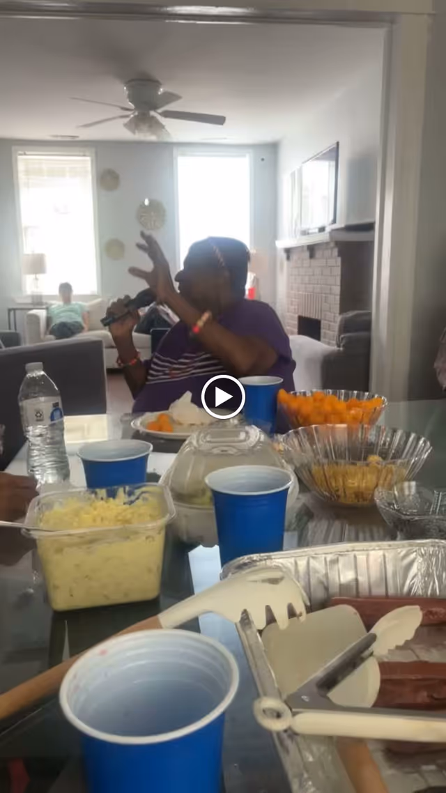 A person holding a microphone and speaking in a living room setting with a table in the foreground containing various food items, blue cups, and utensils. In the background, there are windows, a ceiling fan, and another person sitting on a couch.