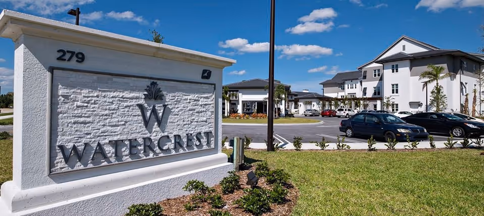 A stone entrance sign reading 'Watercrest' with the senior living complex buildings and parked cars behind it under a blue sky.
