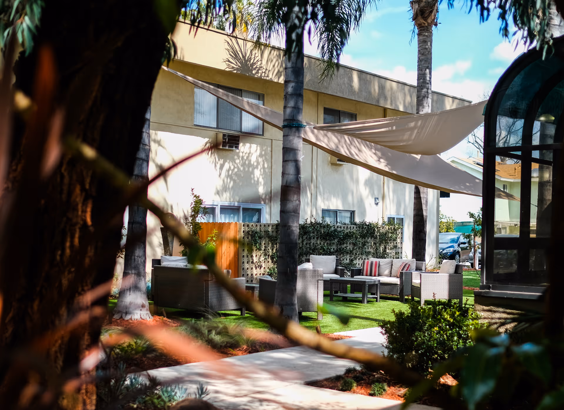 Outdoor seating area at The Gardens at Park Balboa with cushioned chairs and sofas arranged around a coffee table under beige shade sails, surrounded by palm trees and greenery with a building in the background.