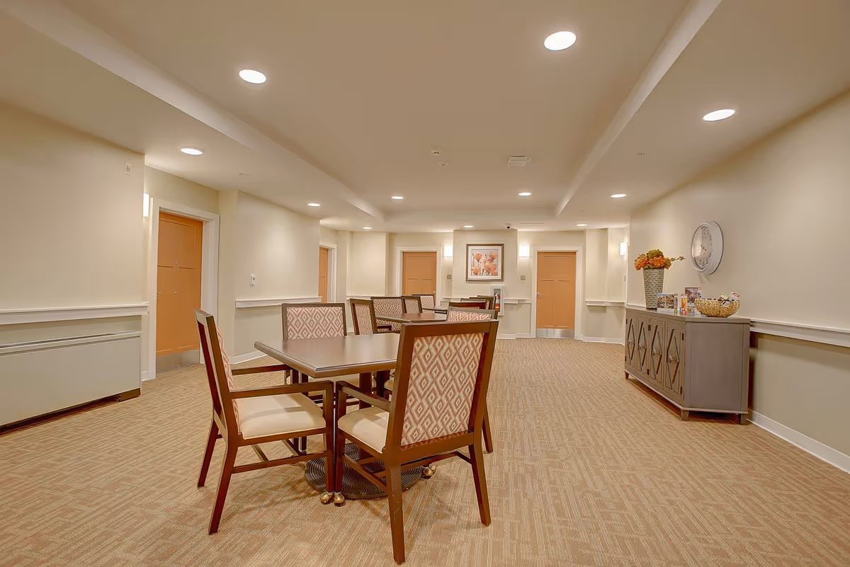 Well-lit communal dining area with a table and chairs, a sideboard, and hallway doors in a senior living facility.