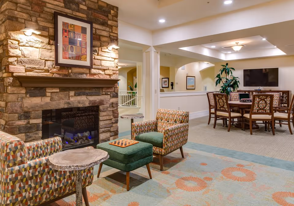 Cozy community lounge with a stone fireplace, patterned armchairs and ottoman, and a table with chairs and TV in the background.