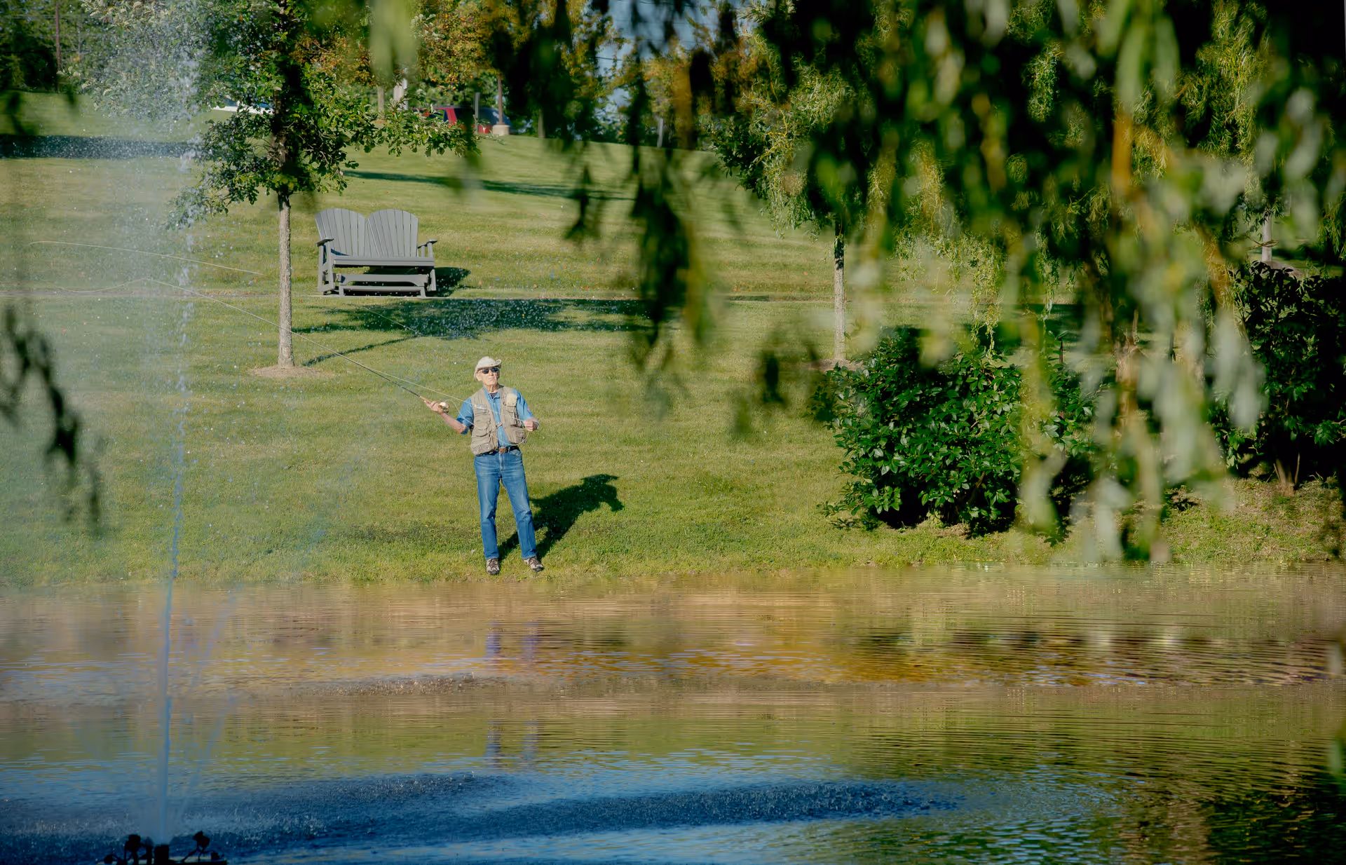 A person fishing by a pond with a water fountain spraying water. The scene includes green grass, trees, bushes, and a wooden bench on a sunny day.
