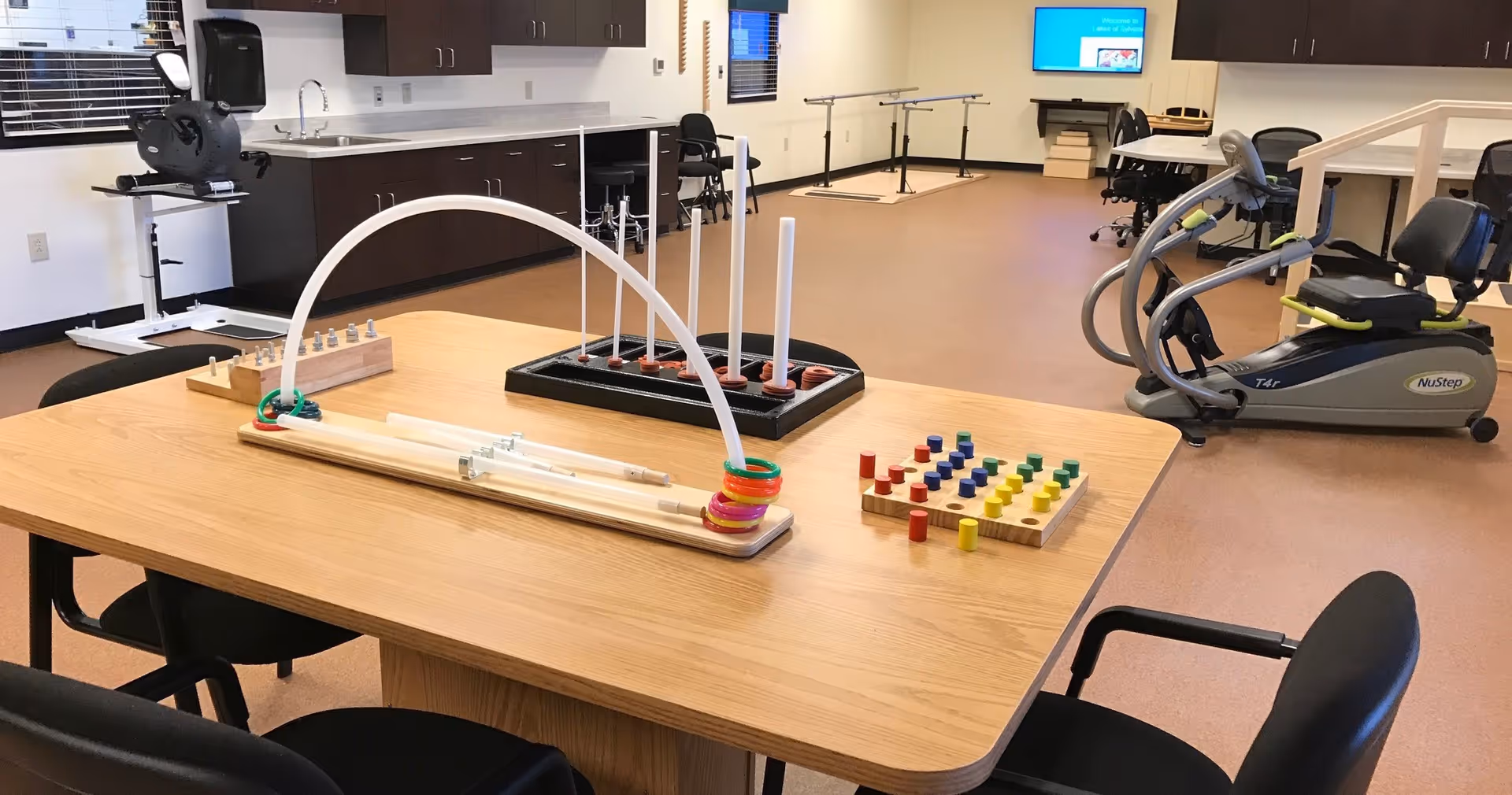Interior therapy/activity room with a wooden table holding peg and ring motor-skills toys, surrounded by chairs and exercise equipment.