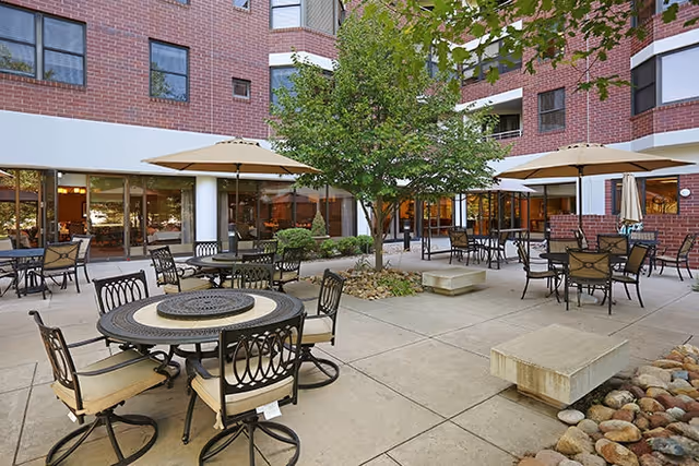 Outdoor patio area at Brookdale Meridian Englewood with multiple round tables and chairs, beige umbrellas providing shade, a tree in the center surrounded by rocks, and a brick building with large windows in the background.