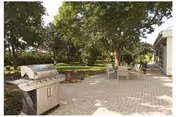 Outdoor patio area with a stainless steel grill on the left, a table with chairs in the center, and large trees providing shade in the background. The patio is paved with bricks and is adjacent to a building on the right.