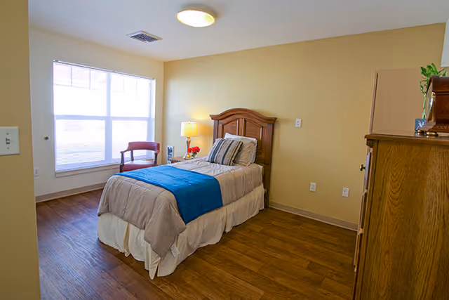 A single bed with beige and blue bedding in a well-lit bedroom with wooden flooring. There is a wooden headboard, a bedside table with a lamp and flowers, a wooden chair near a large window with blinds, and a wooden dresser on the right side.