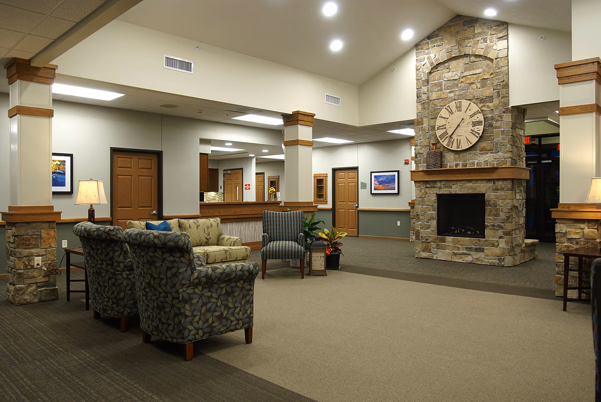 A spacious senior living facility common area with comfortable seating including patterned armchairs and a floral sofa. The room features a large stone fireplace with a rustic clock above it. The walls are painted in neutral tones with wooden trim accents. There are framed pictures on the walls and lamps providing warm lighting. The area has a carpeted floor and an open layout with visible doorways and a reception or kitchen counter in the background.