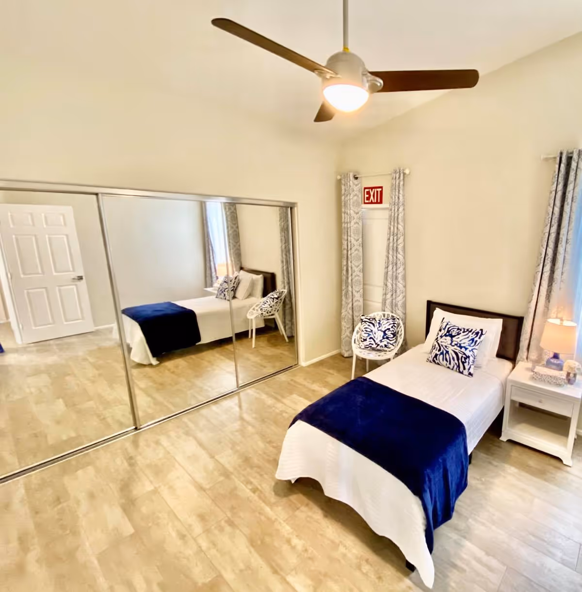 A bright, clean bedroom with a single bed covered in white bedding and a dark blue throw blanket. There is a white nightstand with a lamp and decorative items next to the bed. A white chair with a blue patterned cushion is placed near the bed. The room features a large mirrored closet door, light-colored flooring, and light-colored walls with patterned curtains. A ceiling fan with a light is turned on, and an exit sign is visible above a door in the corner.