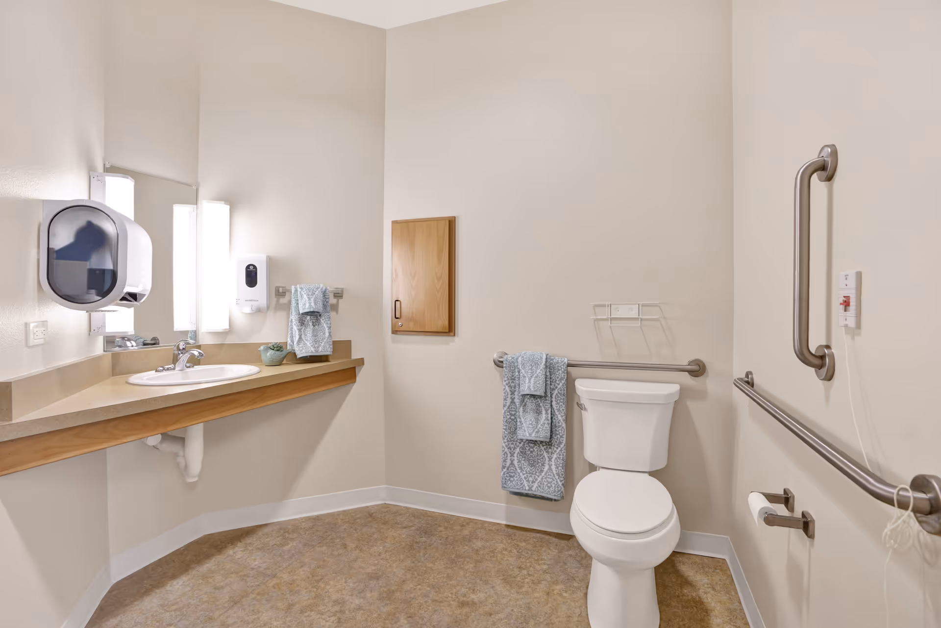 A clean and accessible bathroom with beige walls and flooring. The room features a white toilet with grab bars on both sides, a wall-mounted sink with a countertop, a mirror above the sink, a paper towel dispenser, a soap dispenser, and neatly folded blue patterned towels hanging on a towel rack and grab bar.