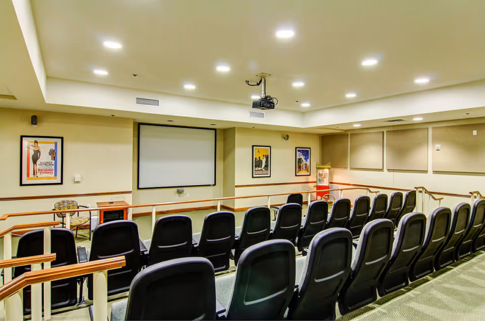 A small theater room with two rows of black cushioned seats facing a large white projection screen. The room has beige walls with framed movie posters and a popcorn machine in the corner. Ceiling lights illuminate the space evenly.