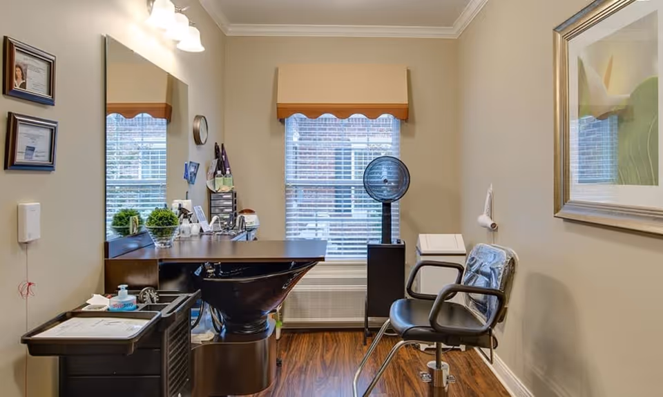 Interior of a senior living facility hair salon with a black salon chair, a black hair washing sink, a rolling cart with supplies, a large mirror, two windows with blinds and valances, a standing hair dryer, and framed certificates and artwork on the walls.