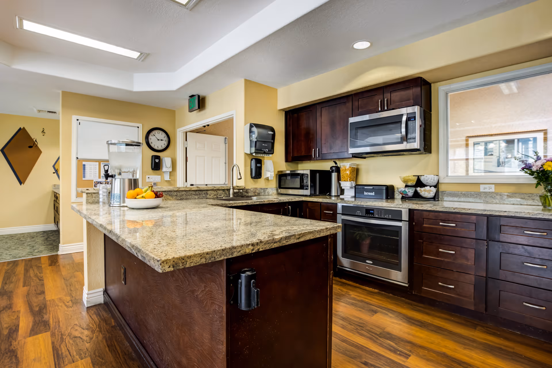 A modern kitchen area with dark wood cabinets, granite countertops, and stainless steel appliances including a microwave and oven. There is a large kitchen island with a water dispenser and a bowl of fruit on it. The floor is wooden, and the walls are painted light yellow. A clock and some wall decorations are visible in the background.