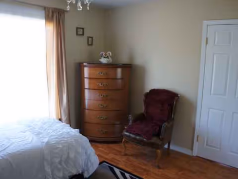 A cozy retirement home bedroom featuring a bed, tall dresser, and burgundy chair.