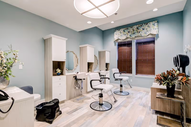 A bright and clean salon room with three white salon chairs in front of mirrors and sinks. The room has light wood flooring, soft blue walls, a large window with wooden blinds and a floral valance, and decorative plants on the counters.