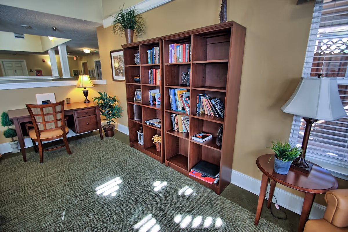 Interior room with a wooden bookshelf filled with books and decorative items, a wooden desk with a chair and a lamp, a potted plant, and a small round table with a lamp and a plant near a window with blinds.