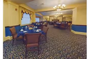 A spacious dining room with multiple round tables covered with blue tablecloths, each set with white cups and saucers. The room features patterned carpet flooring, yellow walls with blue wainscoting, and windows with valances. There are chandeliers hanging from the ceiling and a fireplace in the background.
