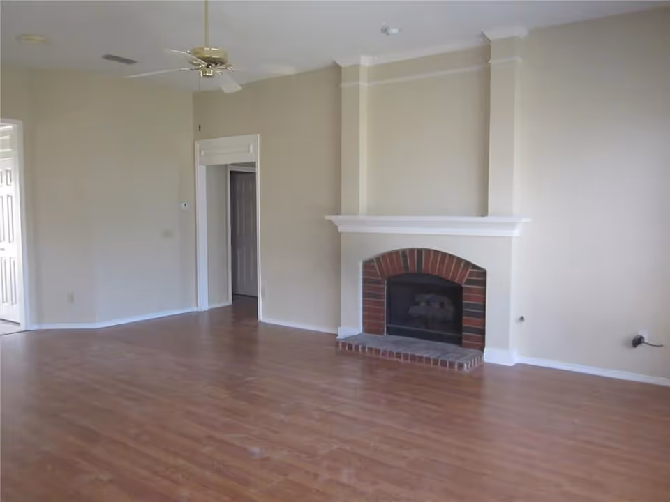 Empty living room with hardwood floors, a brick fireplace and white mantle, a ceiling fan, and a doorway to adjacent rooms.