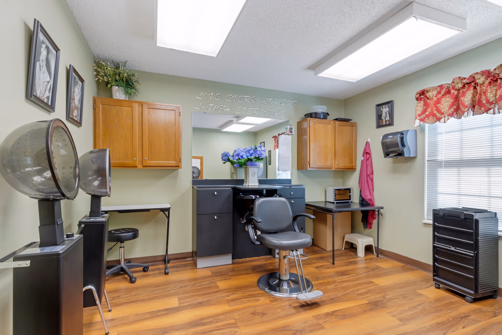 Interior of a hair salon room with a salon chair in front of a black counter with drawers, two wooden wall cabinets, a hair dryer station, a window with red floral valance, and framed pictures on the walls. The room has wood flooring and light green walls.