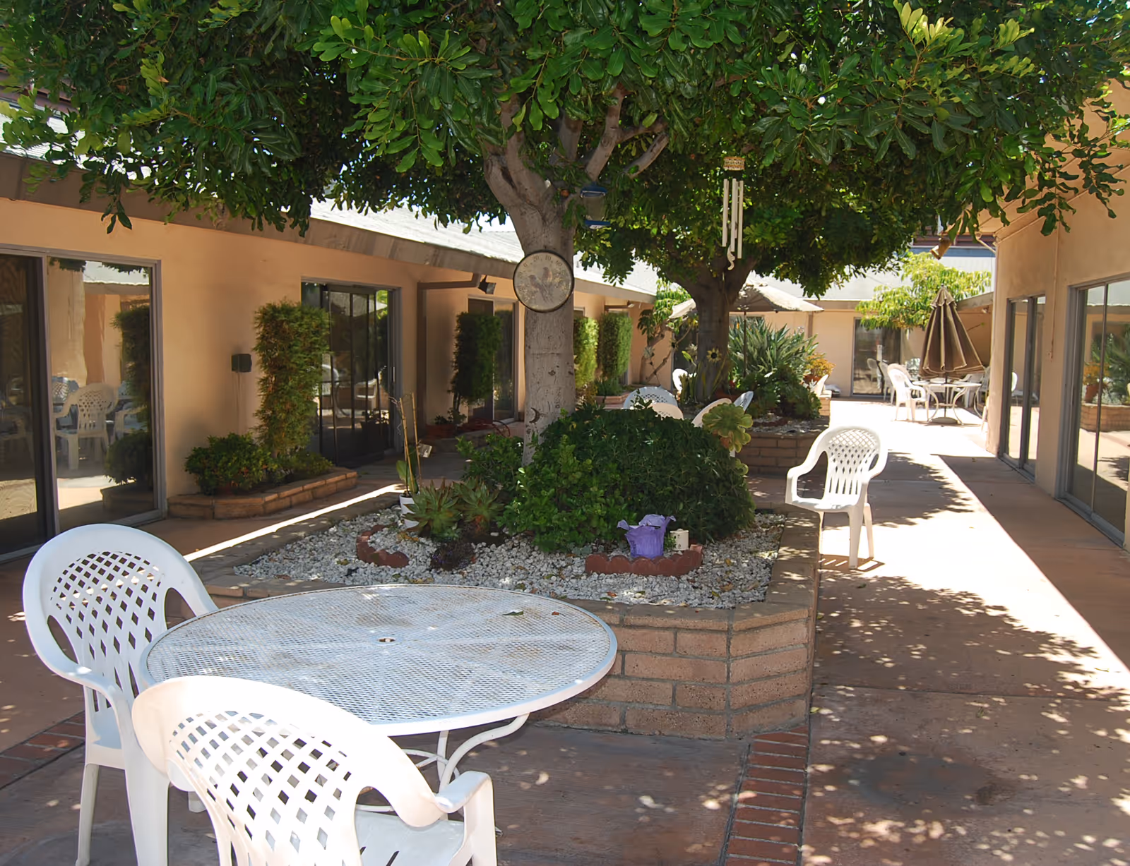 Outdoor courtyard area with white plastic chairs and a round metal table. There are trees providing shade and planters with greenery and small rocks. The courtyard is surrounded by building walls with sliding glass doors.