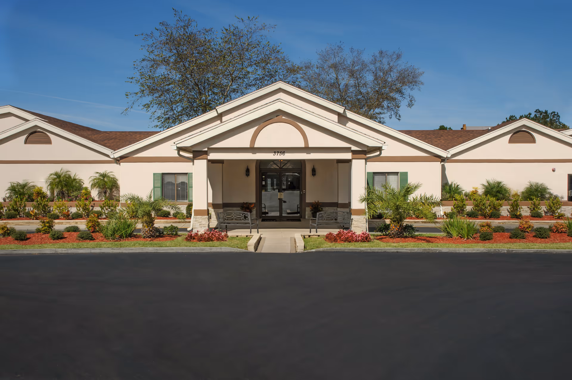 Front entrance of a single-story senior care building with a covered portico, benches, and landscaped flower beds.