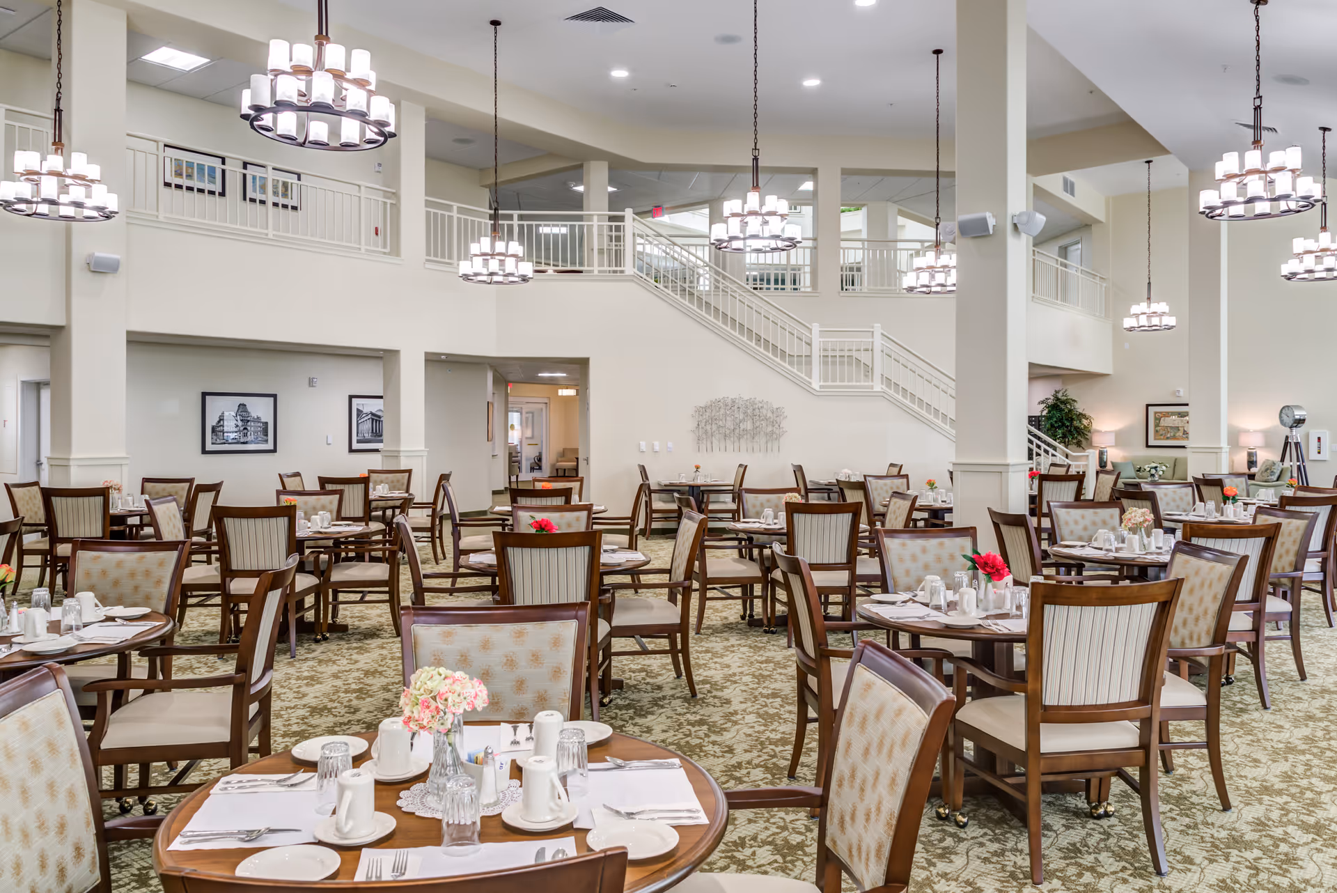 Large elegant dining room with many set tables and chairs, chandeliers, and an upstairs balcony with a staircase.