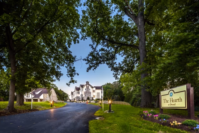 A paved driveway leading to a large, multi-story stone and white building surrounded by green trees and grass. On the right side of the driveway, there is a sign that reads 'The Hearth at Drexel' with flowers planted around its base. The scene is set under a clear blue sky.