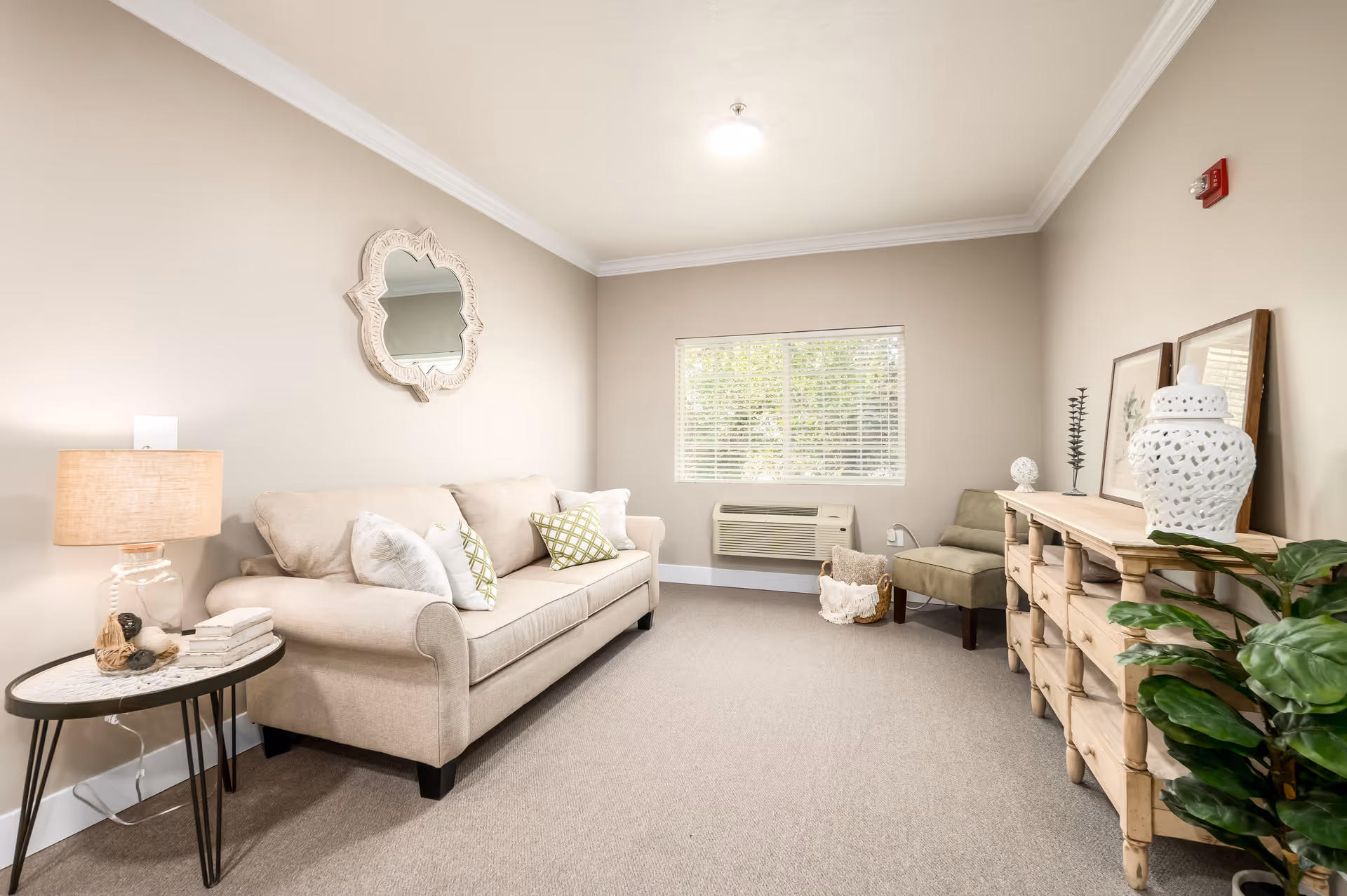 A cozy living room with a beige sofa adorned with patterned and plain cushions, a round side table with a lamp and books, a decorative mirror on the wall, a window with blinds, an air conditioning unit below the window, a green armchair, a wooden console table with decorative items, and a potted plant in the corner.