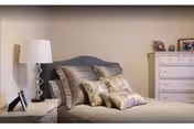 A neatly made bedroom with a bed topped by decorative pillows, a bedside lamp and nightstand, and a dresser with framed photos.